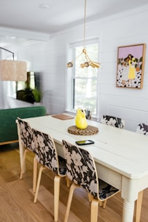 A modern dining room with a white table surrounded by four chairs with floral-patterned backrests. The table has a yellow pitcher, a black smartphone, and a notebook on it. There is a green sofa in the background, a woven pendant lamp, and a plant near the window. The wall features a painting of people with flowers in a yellow background. The room has wooden flooring and white shiplap walls.
