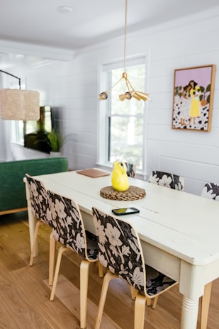 A modern dining room with a white table surrounded by four chairs with floral-patterned backrests. The table has a yellow pitcher, a black smartphone, and a notebook on it. There is a green sofa in the background, a woven pendant lamp, and a plant near the window. The wall features a painting of people with flowers in a yellow background. The room has wooden flooring and white shiplap walls.