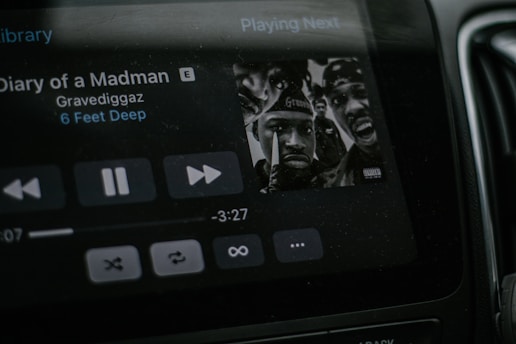 A car stereo display shows a music player screen featuring the album art for a song titled 'Diary of a Madman' by Gravediggaz from the album '6 Feet Deep'. The screen includes the play, pause, and skip buttons, as well as track duration details at the bottom. The album art prominently features several faces.