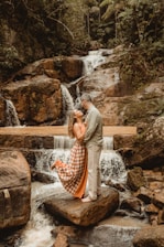 A romantic scene with a couple standing on rocks in front of a cascading waterfall, surrounded by lush greenery. The woman is wearing a flowing checkered dress, while the man is in casual attire, embracing each other affectionately.
