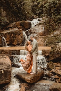 A romantic scene with a couple standing on rocks in front of a cascading waterfall, surrounded by lush greenery. The woman is wearing a flowing checkered dress, while the man is in casual attire, embracing each other affectionately.