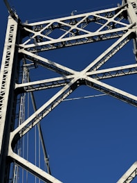 A close-up view of a steel bridge's structural framework against a clear blue sky. The intricate lattice of beams forms a geometric pattern, highlighting the engineering and design of the bridge.