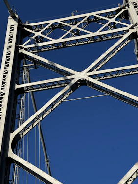 A close-up view of a steel bridge's structural framework against a clear blue sky. The intricate lattice of beams forms a geometric pattern, highlighting the engineering and design of the bridge.