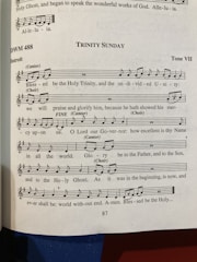 A page from a music book featuring a religious hymn titled 'Trinity Sunday' with sheet music and lyrics. The page number is 87, and the hymn is set in Tone VII. There are notations for cantor and choir parts, with musical notes and lyrics including religious themes.