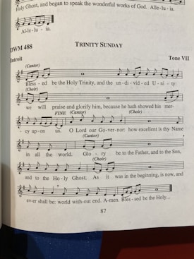 A page from a music book featuring a religious hymn titled 'Trinity Sunday' with sheet music and lyrics. The page number is 87, and the hymn is set in Tone VII. There are notations for cantor and choir parts, with musical notes and lyrics including religious themes.