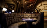 A futuristic, sci-fi setting featuring a circular seating booth with cushioned walls and a central table. The table has illuminated controls and buttons. Surrounding the area are pipes, panels, and electronic components that suggest a spacecraft or advanced technology environment.