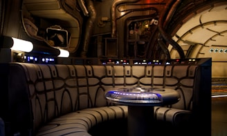 Interior de nave espacial com cabine iluminada, assentos estofados e mesa central com luz azul.