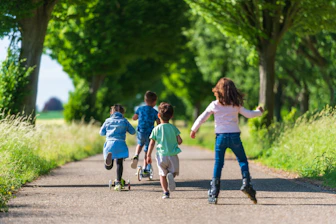 Four children on scooters and rollerblades moving along a tree-lined path in summer