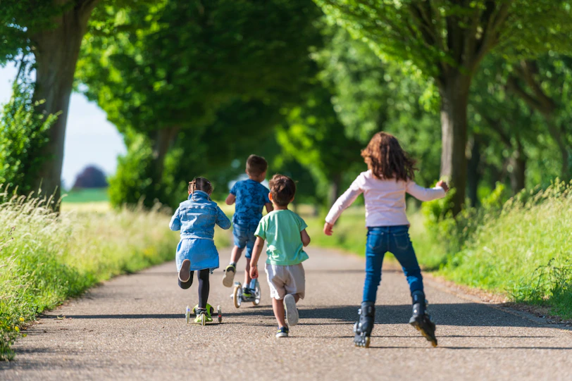 Four children on scooters and rollerblades moving along a tree-lined path in summer