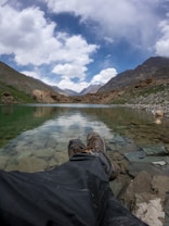 A serene landscape with a tranquil lake reflecting the sky, surrounded by rocky terrain and distant mountains under a partly cloudy sky. The foreground shows someone relaxing with legs stretched out, wearing hiking boots and dark pants, enhancing the sense of personal connection to nature.