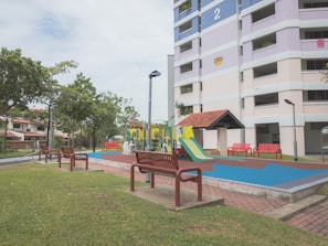 A residential area with a playground featuring colorful slides and climbing structures. Surrounding the playground are several red benches and green grass. A multi-story apartment building with pastel hues is visible in the background alongside well-maintained trees and street lamps.