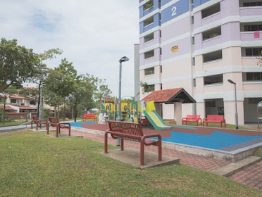 A residential area with a playground featuring colorful slides and climbing structures. Surrounding the playground are several red benches and green grass. A multi-story apartment building with pastel hues is visible in the background alongside well-maintained trees and street lamps.