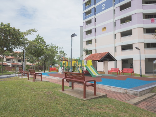 A residential area with a playground featuring colorful slides and climbing structures. Surrounding the playground are several red benches and green grass. A multi-story apartment building with pastel hues is visible in the background alongside well-maintained trees and street lamps.