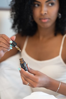 A woman with curly hair is holding a small bottle of oil and using a dropper to extract some of it. She appears to be in a relaxed setting, possibly at home, wearing a white camisole-like top. Her nails are painted light blue, and she wears a ring and a bracelet.
