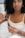 A woman with curly hair is holding a small bottle of oil and using a dropper to extract some of it. She appears to be in a relaxed setting, possibly at home, wearing a white camisole-like top. Her nails are painted light blue, and she wears a ring and a bracelet.