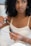 A woman with curly hair is holding a small bottle of oil and using a dropper to extract some of it. She appears to be in a relaxed setting, possibly at home, wearing a white camisole-like top. Her nails are painted light blue, and she wears a ring and a bracelet.