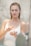 A person in a white tank top is applying a cream or lotion with a focused expression, standing in a softly lit environment that appears to be a bathroom. The background is blurred, enhancing the serene and intimate atmosphere.