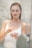 A person in a white tank top is applying a cream or lotion with a focused expression, standing in a softly lit environment that appears to be a bathroom. The background is blurred, enhancing the serene and intimate atmosphere.