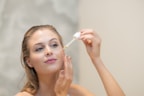 A woman is applying skincare serum to her face using a dropper. She appears relaxed and focused, with her hair pulled back, allowing the focus to be on her facial routine. The background is a soft, neutral tone.