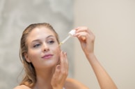 A woman is applying skincare serum to her face using a dropper. She appears relaxed and focused, with her hair pulled back, allowing the focus to be on her facial routine. The background is a soft, neutral tone.