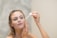 A woman is applying skincare serum to her face using a dropper. She appears relaxed and focused, with her hair pulled back, allowing the focus to be on her facial routine. The background is a soft, neutral tone.