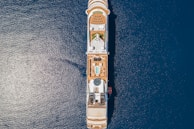 An overhead view of a large cruise ship sailing on deep blue ocean waters. The ship's top deck features rows of lounge chairs, a swimming pool, a circular glass dome structure, and various recreational areas. The symmetry and layout of the ship are clearly visible from this angle.