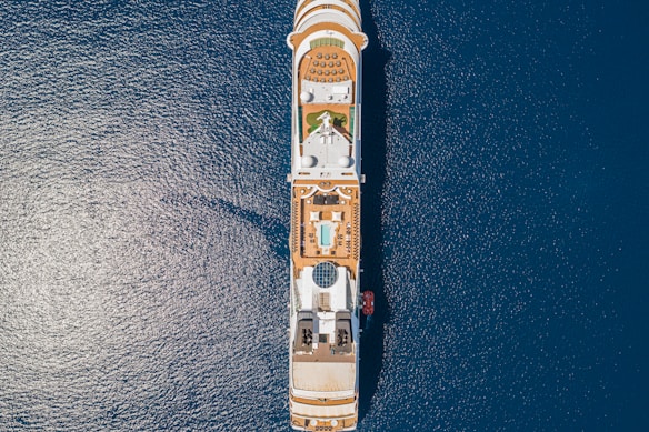 An overhead view of a large cruise ship sailing on deep blue ocean waters. The ship's top deck features rows of lounge chairs, a swimming pool, a circular glass dome structure, and various recreational areas. The symmetry and layout of the ship are clearly visible from this angle.