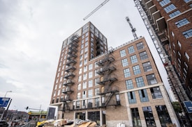 A tall, multi-story brick building under construction, featuring large windows and external fire escape staircases. Cranes are visible in the background, and construction materials are scattered in the foreground. The sky is overcast, adding a sense of industrial progress to the scene.
