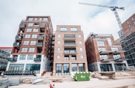 A construction site featuring several multi-story brick buildings with large windows and balconies. A crane is visible on the right side of the image, indicating ongoing construction activity. Stacks of wooden planks and other construction materials are scattered in the foreground. A green dumpster is placed in front of the buildings.