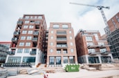 A construction site featuring several multi-story brick buildings with large windows and balconies. A crane is visible on the right side of the image, indicating ongoing construction activity. Stacks of wooden planks and other construction materials are scattered in the foreground. A green dumpster is placed in front of the buildings.