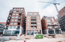 A construction site featuring several multi-story brick buildings with large windows and balconies. A crane is visible on the right side of the image, indicating ongoing construction activity. Stacks of wooden planks and other construction materials are scattered in the foreground. A green dumpster is placed in front of the buildings.
