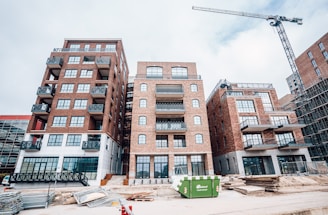 A construction site featuring several multi-story brick buildings with large windows and balconies. A crane is visible on the right side of the image, indicating ongoing construction activity. Stacks of wooden planks and other construction materials are scattered in the foreground. A green dumpster is placed in front of the buildings.
