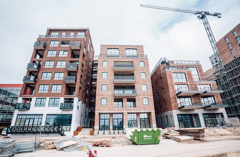 A construction site featuring several multi-story brick buildings with large windows and balconies. A crane is visible on the right side of the image, indicating ongoing construction activity. Stacks of wooden planks and other construction materials are scattered in the foreground. A green dumpster is placed in front of the buildings.