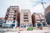A construction site featuring several multi-story brick buildings with large windows and balconies. A crane is visible on the right side of the image, indicating ongoing construction activity. Stacks of wooden planks and other construction materials are scattered in the foreground. A green dumpster is placed in front of the buildings.