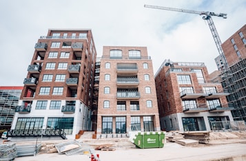 A construction site featuring several multi-story brick buildings with large windows and balconies. A crane is visible on the right side of the image, indicating ongoing construction activity. Stacks of wooden planks and other construction materials are scattered in the foreground. A green dumpster is placed in front of the buildings.