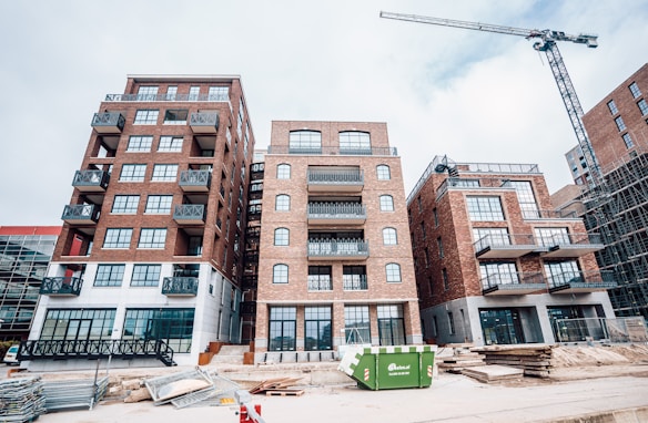 A construction site featuring several multi-story brick buildings with large windows and balconies. A crane is visible on the right side of the image, indicating ongoing construction activity. Stacks of wooden planks and other construction materials are scattered in the foreground. A green dumpster is placed in front of the buildings.