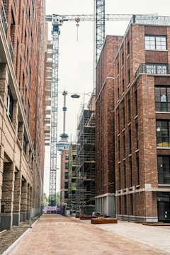 A construction site between tall brick buildings with a crane in the center. Scaffolding is visible on one of the buildings, indicating ongoing construction work. The scene is quite industrial, with a dirt pathway leading into the distance.
