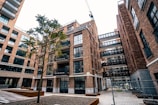 An urban scene featuring a modern apartment complex with multiple brick buildings. The architecture includes large glass windows and several balconies. A lone tree stands in the courtyard area, and a crane is visible in the background, indicating ongoing construction. The area is fenced, suggesting construction work.