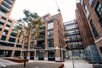 An urban scene featuring a modern apartment complex with multiple brick buildings. The architecture includes large glass windows and several balconies. A lone tree stands in the courtyard area, and a crane is visible in the background, indicating ongoing construction. The area is fenced, suggesting construction work.