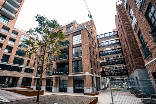An urban scene featuring a modern apartment complex with multiple brick buildings. The architecture includes large glass windows and several balconies. A lone tree stands in the courtyard area, and a crane is visible in the background, indicating ongoing construction. The area is fenced, suggesting construction work.