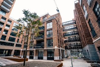 An urban scene featuring a modern apartment complex with multiple brick buildings. The architecture includes large glass windows and several balconies. A lone tree stands in the courtyard area, and a crane is visible in the background, indicating ongoing construction. The area is fenced, suggesting construction work.