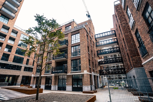 An urban scene featuring a modern apartment complex with multiple brick buildings. The architecture includes large glass windows and several balconies. A lone tree stands in the courtyard area, and a crane is visible in the background, indicating ongoing construction. The area is fenced, suggesting construction work.