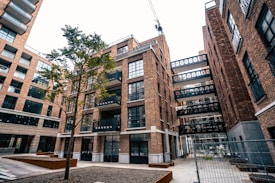 An urban scene featuring a modern apartment complex with multiple brick buildings. The architecture includes large glass windows and several balconies. A lone tree stands in the courtyard area, and a crane is visible in the background, indicating ongoing construction. The area is fenced, suggesting construction work.