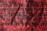 A wrinkled poster with overlapping, bold black text on a red background. The text appears distressed and chaotic, with phrases such as 'HUSTLE OF THE SYSTEM' and other fragmented words, suggesting a theme of defiance or chaos.