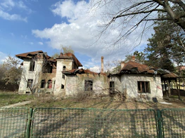 A worn-down house with visible damage, surrounded by greenery under a bright sky.