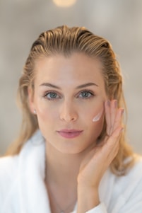 A person with light hair and blue eyes applies a cream to their cheek using their hand. The person wears a white robe and appears to be engaged in a skincare routine. The background is blurred, emphasizing the subject's facial features and skincare application.
