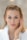 A person with light hair and blue eyes applies a cream to their cheek using their hand. The person wears a white robe and appears to be engaged in a skincare routine. The background is blurred, emphasizing the subject's facial features and skincare application.