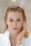 A person with light hair and blue eyes applies a cream to their cheek using their hand. The person wears a white robe and appears to be engaged in a skincare routine. The background is blurred, emphasizing the subject's facial features and skincare application.