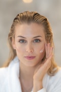 A person with light hair and blue eyes applies a cream to their cheek using their hand. The person wears a white robe and appears to be engaged in a skincare routine. The background is blurred, emphasizing the subject's facial features and skincare application.