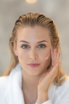 A person with light hair and blue eyes applies a cream to their cheek using their hand. The person wears a white robe and appears to be engaged in a skincare routine. The background is blurred, emphasizing the subject's facial features and skincare application.